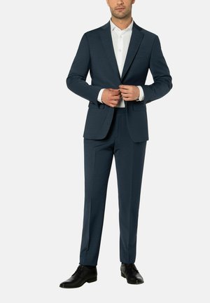 Man die een donkerblauwe colbert over een wit overhemd knoopt, met bijpassende broek en zwarte schoenen, staat op een witte achtergrond.