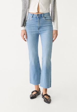 Vrouw draagt lichtblauwe, hoog uitgesneden, cropped flare jeans met zwarte puntige ballerina's versierd met zilveren studs.