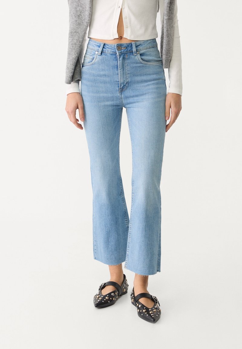 Une femme porte un jean flare court taille haute bleu clair avec des ballerines noires pointues ornées de clous argentés.