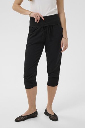 Naine, kes kannab musti lühikesi pükse, millel on nööriga vöökoht, ja musti võrkmaterjalist baleriinikingi, seisab ühevärvilise tausta ees.