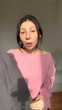 Νέα γυναίκα με σκούρα μαλλιά και ασημένια σκουλαρίκια φοράει ροζ τοπ με ανοιχτούς ώμους, κρατώντας μια διπλωμένη γκρι ζακέτα σε έναν απαλά φωτισμένο χώρο.