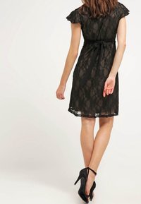 Robe en dentelle noire avec des manches courtes et volantes, une taille ajustée et un lien au dos, avec un ourlet festonné et une texture transparente.
