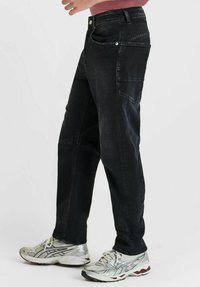 Zij-aanzicht van een persoon in zwarte denimspijkerbroek en witte hardloopschoenen, staande tegen een effen achtergrond.