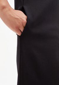 Vêtement noir avec une poche latérale et une main placée à l'intérieur. La surface du tissu semble lisse avec une texture douce.