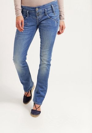 Femme portant un jean slim bleu avec une fermeture triple boutons et des espadrilles plates noires, debout devant un fond blanc uni.