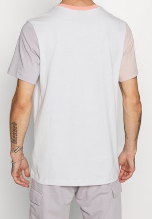 Mann trägt ein weißes T-Shirt mit pastellroten und lila Ärmeln und steht vor einem schlichten hellen Hintergrund, von hinten sichtbar.