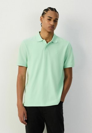 Fiatal férfi fonott hajjal, világoszöld rövid ujjú galléros pólót és fekete nadrágot visel, egyszínű fehér háttér előtt áll.