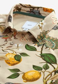 Chemise beige avec imprimé botanique comprenant des citrons, des feuilles vertes, des fleurs et un motif de paon, pliée avec col et boutons visibles.