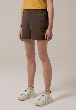 Persona vistiendo pantalones cortos cargo marrones con bolsillo y zapatos deportivos blancos, de pie contra un fondo liso.