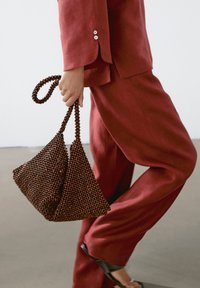 Sac à main marron en perles avec une forme triangulaire et une anse tressée, associé à un costume en lin rouge. Texture lisse et motif de perles complexe.