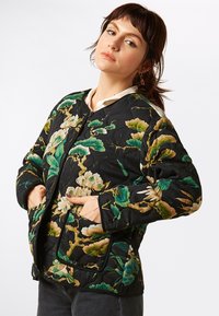 Gequilted zwarte jas met bloemenpatronen in groen, crème en goud. Beschikt over twee zijzakken en een ronde halslijn met knoopsluiting.