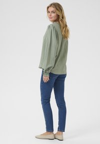 Blouse verte à manches longues avec des épaules froncées et des détails aux poignets, associée à un jean skinny bleu et des ballerines beiges, vue de dos.