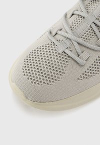 Light gray athletic shoe featuring breathable mesh upper, textured pattern, and flat laces. The sole is lightly colored and rounded at the toe.