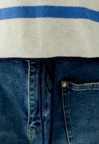 Strickpullover in Creme mit blauen Streifen am Saum, kombiniert mit dunkelblauen Jeans, die eine sichtbare Tasche und einen Kordelzug haben.