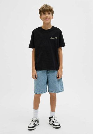 Dječak stoji, nosi crnu majicu, svijetloplave traper kratke hlače, bijele čarape i crno-bijele tenisice na bijeloj pozadini.