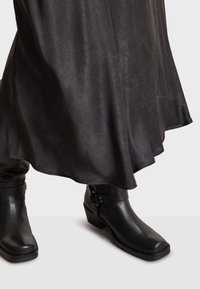 Bottines en cuir noir avec un bout carré, un petit talon bloc et un détail de boucle décoratif ; portées avec une jupe fluide et texturée de couleur sombre.