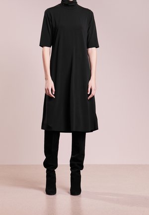 Persona con un vestido negro hasta la rodilla y mangas hasta el codo, combinado con botas negras hasta la rodilla, de pie frente a un fondo neutral.