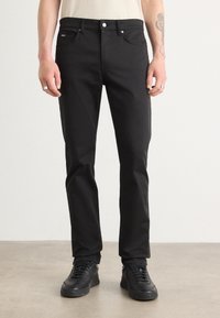 Svarta jeans i denim med rak passform, utrustade med framlådor och knappgylf. Bärs med svarta sneakers, mot en neutral bakgrund.