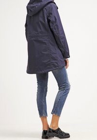 Navy waterproof jacket with a hood, elastic cuffs, and a thigh-length cut, paired with fitted light blue jeans and black shoes.
