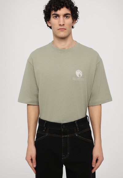 Camiseta holgada de color verde claro en algodón con mangas cortas, escote redondo y un pequeño diseño de logo impreso en el pecho. Combinada con jeans negros.