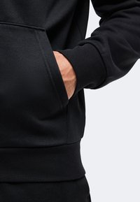Main dans la poche avant d'un sweat-shirt noir à manches longues avec poignets et ourlet côtelés.