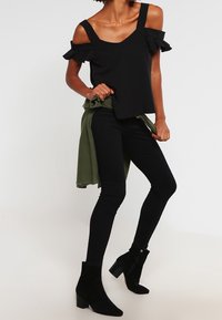 Top nero senza spalline con dettagli a volant, abbinato a jeans skinny neri e stivaletti neri, stratificato con un indumento verde legato in vita.