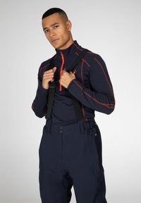 Marineblauwe zip-up top met rode accenten, gecombineerd met marineblauwe broek en zwarte bretels. Glad materiaal, aansluitend ontwerp en functionele hardwaredetails.
