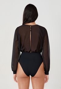 Bodysuit negro con mangas largas transparentes y cuello redondo. La parte posterior presenta una abertura en forma de llave con cierre de botón. Diseño de pierna de corte alto.