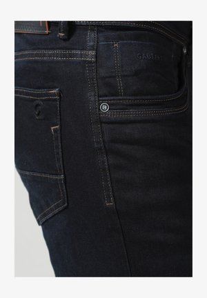 Dunkelblaue Denim-Jeans mit schmaler Passform, auffälliger Naht auf der Gesäßtasche, einem kleinen Markenakzent und einer glatten Textur.