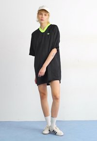 Svart oversized t-shirt med grön krage, korta ärmar, logotyp på bröstet. Modellen bär vit keps och strumpor med svarta ränder och svarta skor.
