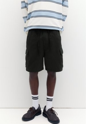 Person iført sorte cargoshorts, hvide sokker med sorte striber, sorte sko og en blå- og hvidstribet langærmet skjorte.