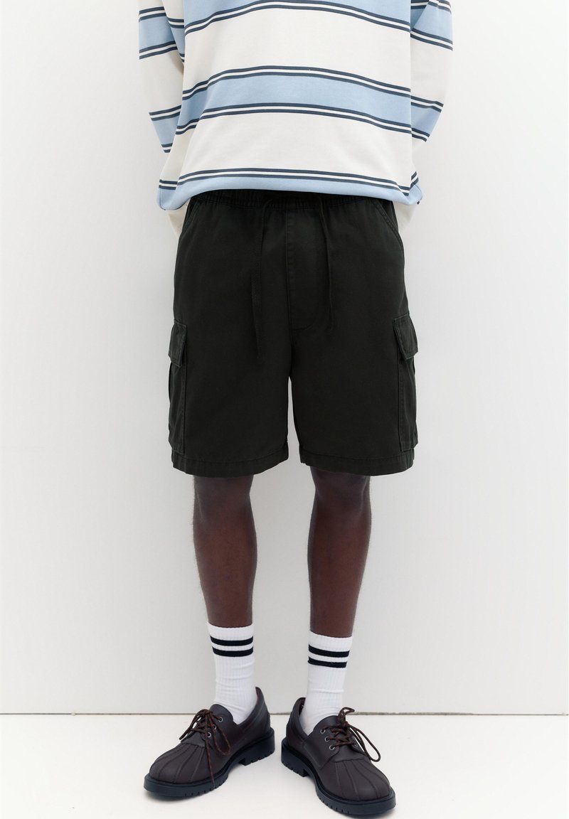 Personne portant un short cargo noir, des chaussettes blanches avec des rayures noires, des chaussures noires et un t-shirt à manches longues rayé bleu et blanc.