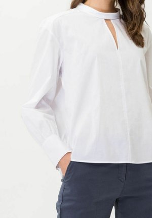 Woman wearing a long-sleeve white blouse with a front keyhole neckline, paired with dark navy pants, hand in pocket.