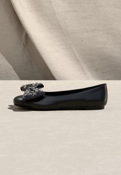 Μαύρο επίπεδο παπούτσι με μεγάλο φιόγκο διακοσμημένο με ασημένια καρφιά στη μύτη, τοποθετημένο σε μπεζ υφασμάτινο φόντο.