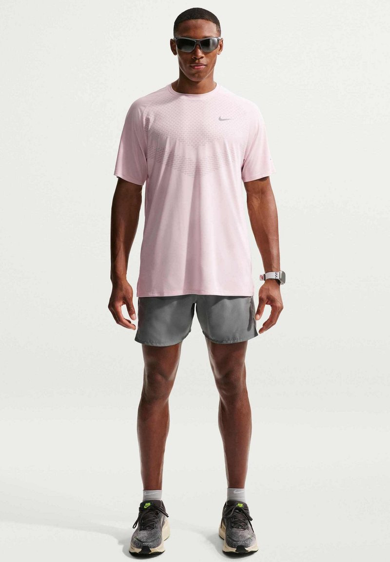 Mann mit Sonnenbrille, pinkem Sportshirt, grauen Shorts und schwarzen Laufschuhen, stehend vor einem einfarbigen hellen Hintergrund.