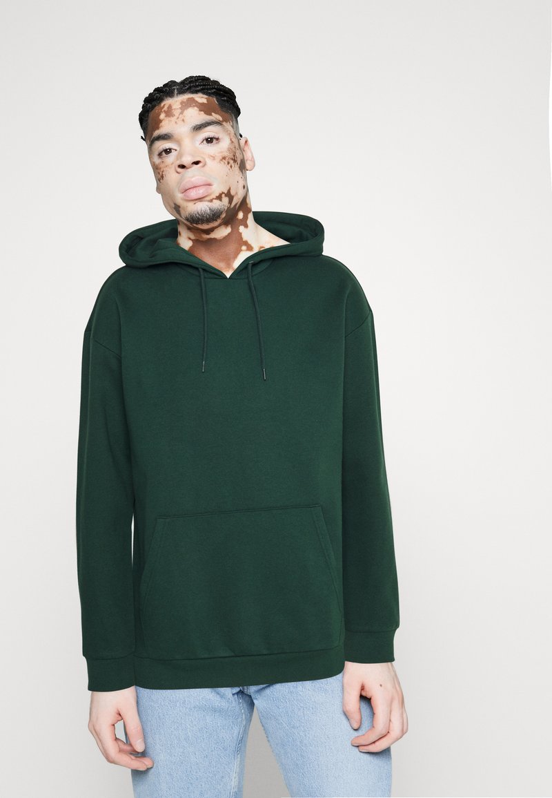 Donkergroen sweatshirt met capuchon, gemaakt van zachte stof. Voorzien van een voorzak, een trekkoordkap en geribbelde boorden; gecombineerd met lichtblauwe spijkerbroek.