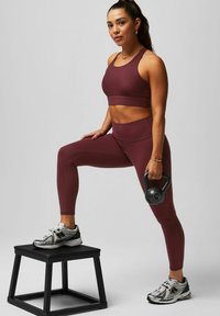 Bordeauxfarbener, sportlicher Cropped-Top und Leggings, enganliegend, mit glatter Textur. Trägt schwarze und silberne Sneaker. Hält eine schwarze Kettlebell.