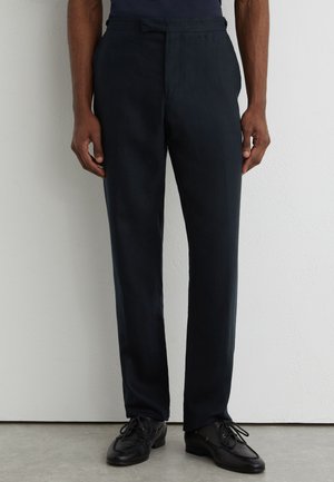 Muž v čiernych na mieru šitých nohaviciach a čiernych kožených spoločenských topánkach stojaci pri bielej stene a šedej podlahe.