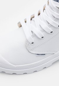 Sneaker bianco con una tomaia sintetica liscia, dettagli texturizzati e lacci bianchi. Presenta un'etichetta con logo e una suola in gomma robusta.