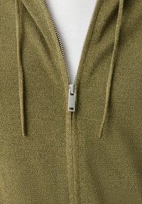 Nahaufnahme eines olivgrünen, gestrickten Zip-Hoodies mit silbernem Reißverschluss, der teilweise geöffnet ist und ein weißes Shirt darunter zeigt.