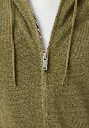 Nahaufnahme eines olivgrünen, gestrickten Zip-Hoodies mit silbernem Reißverschluss, der teilweise geöffnet ist und ein weißes Shirt darunter zeigt.