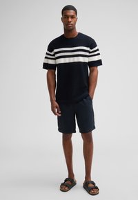 Marineblauer Strickpullover mit weißen und grauen horizontalen Streifen, kombiniert mit dunklen Shorts und schwarzen Sandalen. Das Model steht mit den Händen in den Taschen.