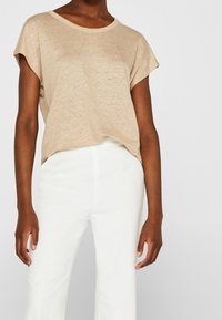 T-shirt a maniche corte di un leggero beige, con scollatura rotonda, abbinato a pantaloni bianchi sartoriali. Il tessuto sembra morbido con una finitura leggermente texturizzata.