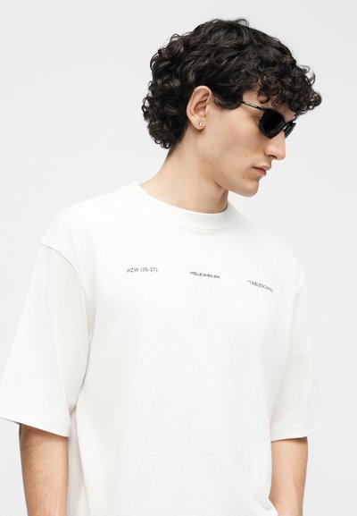 Νέος άνδρας με σγουρά μαλλιά που φοράει μαύρα γυαλιά ηλίου και μία λευκή υπερμεγέθη μπλούζα με μικρό τυπωμένο κείμενο στο στήθος.