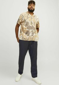 Camisa de manga corta beige con patrones florales y abstractos marrones, combinada con pantalones azul marino oscuros y zapatillas blancas. Diseño simple y ajuste relajado.