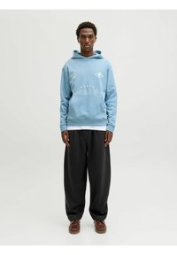 Sudadera con capucha azul con texto gráfico blanco y acentos, combinada con pantalones negros holgados y zapatos de cordones marrones.