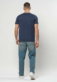 Moški, ki odhaja, nosi temno modro majico, svetlo modre kavbojke, bele superge in srebrno verižico na preprostem ozadju.