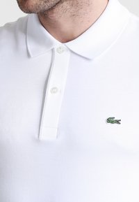 Bela polo majica s klasičnim ovratnikom, ki ima na levem prsnem delu zeleno vezeno logotip krokodila. Narejena iz gladkega blaga, z minimalnimi detajli.
