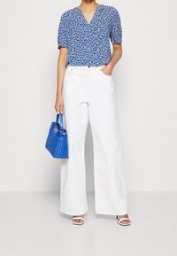 Blouse bleue à manches courtes avec motif abstrait blanc, associée à un pantalon blanc à jambes larges et un sac bleu tissé. Escarpins blancs à bouts ouverts.