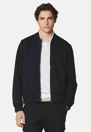 Junger Mann mit kurzen, lockigen braunen Haaren, der eine schwarze Reißverschlussjacke über einem weißen Poloshirt trägt und vor einem einfarbigen Hintergrund steht.